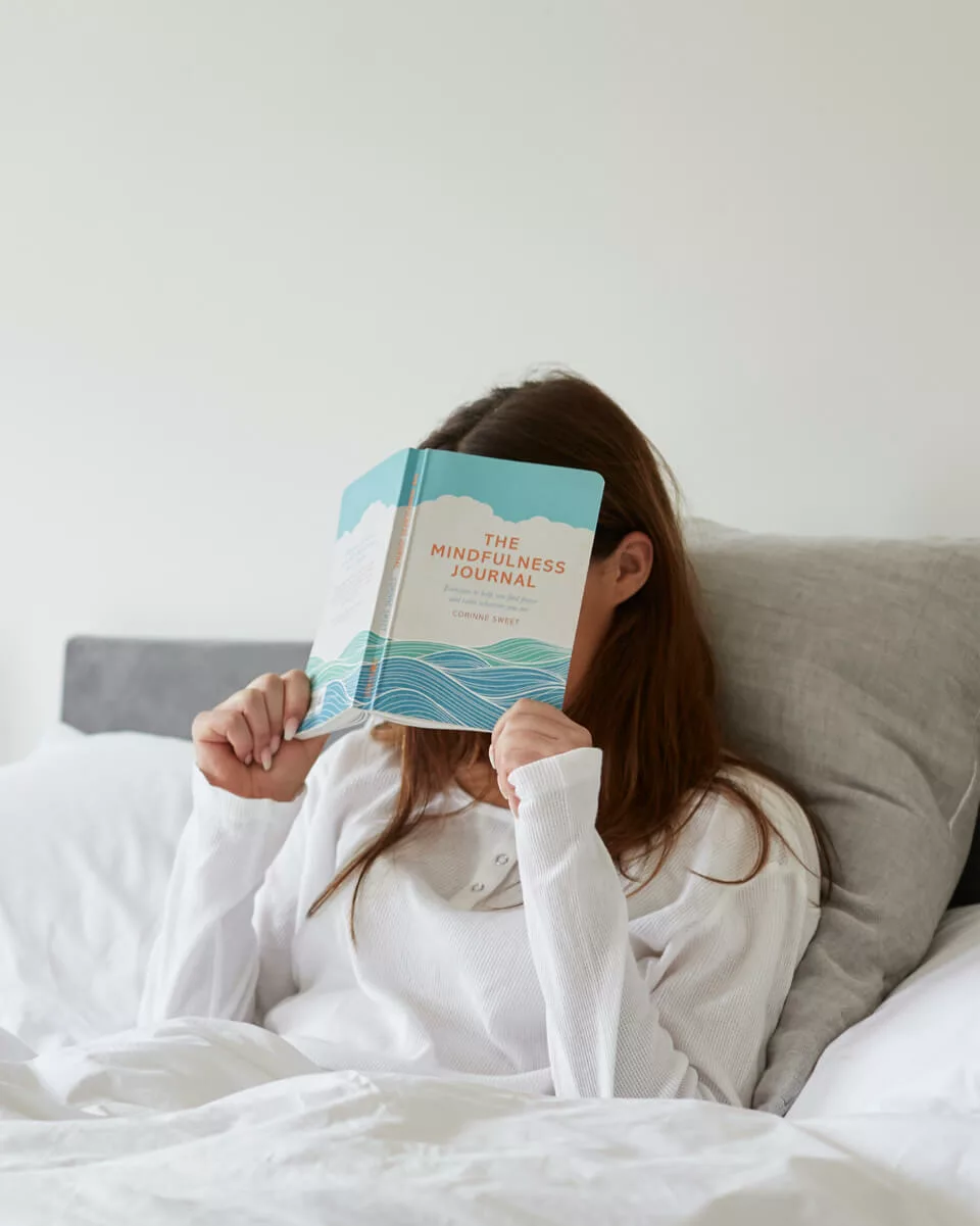 A woman laying in bed wearing a white, long sleeve top with a book over her face